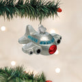 A silver airplane ornament with blue and red details hangs from a green Christmas tree branch, with blurred white holiday lights in the background.