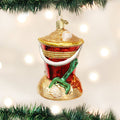A Christmas tree ornament shaped like a red grill on a sand base with seashells and a green shovel, hanging among green branches with blurred white lights in the background.