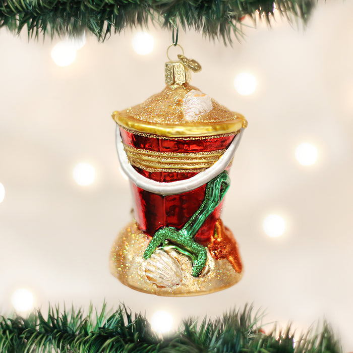 A Christmas tree ornament shaped like a red grill on a sand base with seashells and a green shovel, hanging among green branches with blurred white lights in the background.