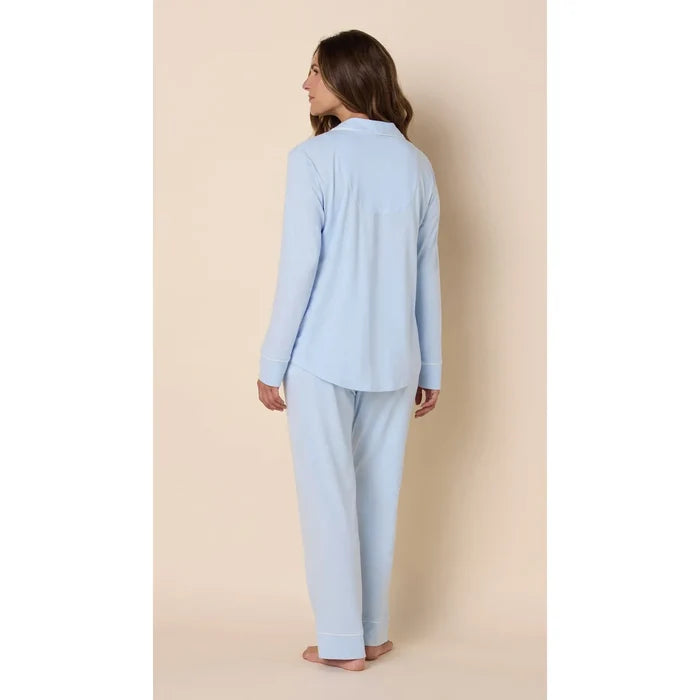 A woman wearing a light blue two-piece pajama set, consisting of a long-sleeve button-up top and matching pants, shown from a rear three-quarter view against a solid beige studio background.