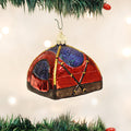 A red and purple tent-shaped Christmas ornament with gold accents hangs from a green tree branch, with blurred white lights in the background.