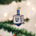 A blue and silver dreidel ornament hangs from a green pine branch, with soft white lights blurred in the background.