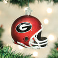A red football helmet ornament with a black G logo hangs on a Christmas tree. The ornament is decorated with white details and surrounded by green branches and blurred holiday lights in the background.