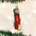 A shiny red Christmas tree ornament shaped like a golf bag with silver clubs and a small golden dog figure at the base hangs from a green tree branch. White holiday lights glow in the blurred background.