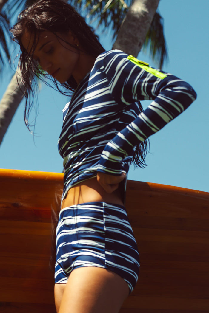 Woman wearing a blue & white striped long sleeve tee and boy short holding a surfboard in a tropical background.