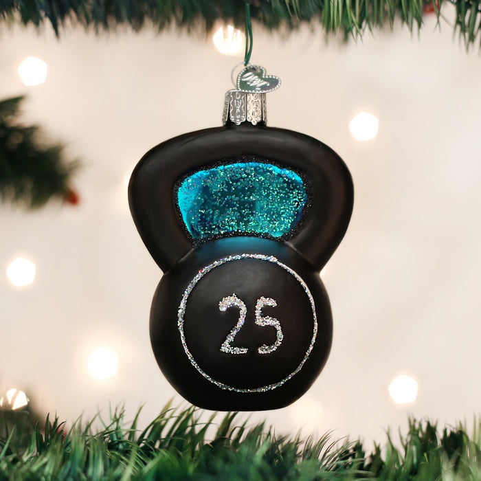 A Christmas tree ornament shaped like a black kettlebell with blue glitter accents and the number 25 written in silver glitter, hanging among green pine branches with white lights in the background.