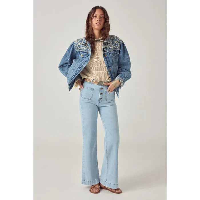 Woman wearing a denim jacket, beige sweater, and light blue jeans on a plain background
