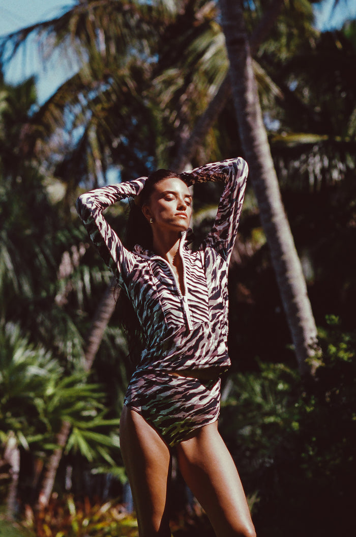 Woman modeling Watskin Mia rashguard and Aria bikini bottoms in Animal Mix against a tropical beach scene with palm trees