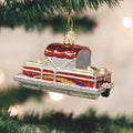 A shiny Christmas ornament shaped like a classic red and silver car hangs from a green pine tree branch. The background is softly blurred with white holiday lights.
