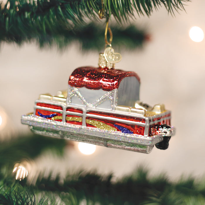 A shiny Christmas ornament shaped like a classic red and silver car hangs from a green pine tree branch. The background is softly blurred with white holiday lights.