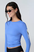 3/4 view of a woman wearing a light blue long-sleeve sun protective tee with side ruching and arm zip pocket and black bikini bottom