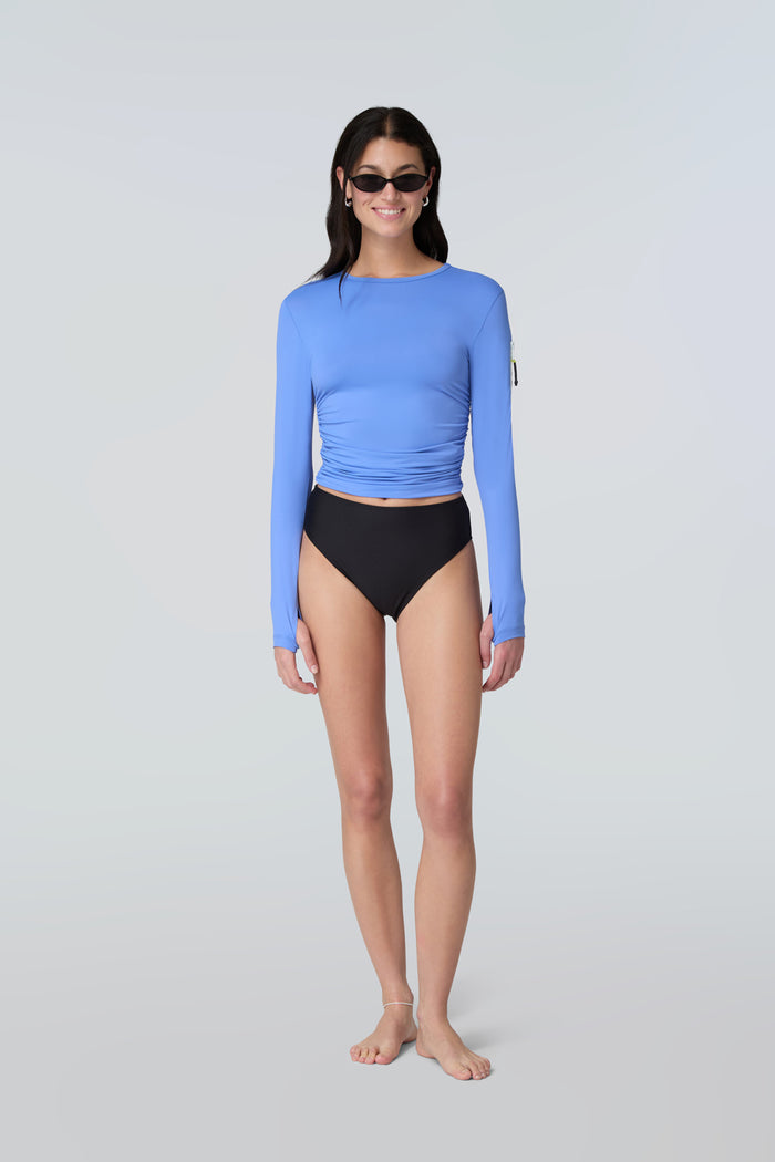 Front view of a woman wearing a light blue long-sleeve sun protective tee with side ruching and arm zip pocket and black bikini bottom