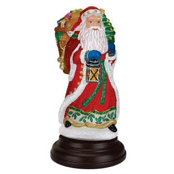 A festive Santa Claus figurine in a red coat holds a lantern and a green Christmas tree, with a sack of gifts on his back, standing on a dark round base.