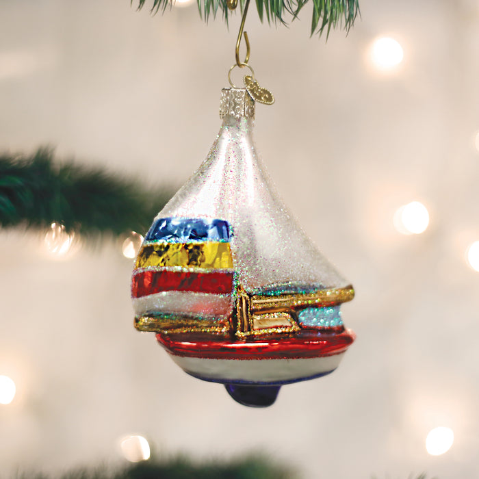 A colorful sailboat-shaped ornament with glitter, hanging from a Christmas tree branch, with soft white lights blurred in the background.