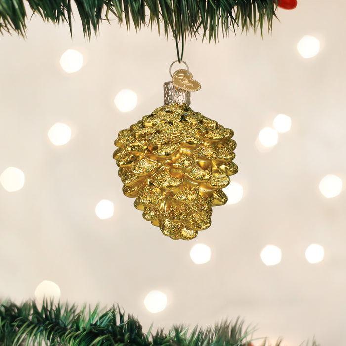 A gold, glittery pine cone ornament hangs from a Christmas tree branch with blurred white holiday lights glowing softly in the background.