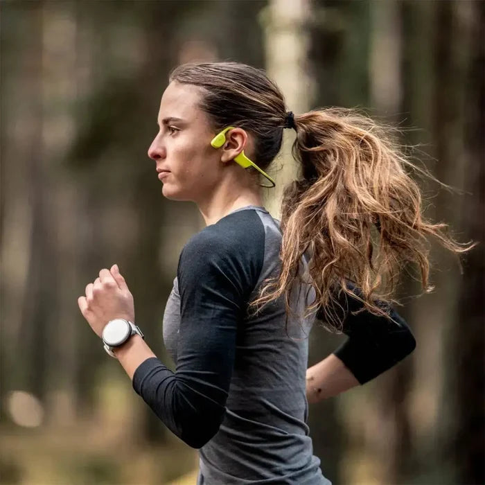 Woman running in the forest wearing Suunto Sonic Open Ear Bone Headphones in yellow, demonstrating their secure fit for active lifestyles.  from  John Bull Clothing