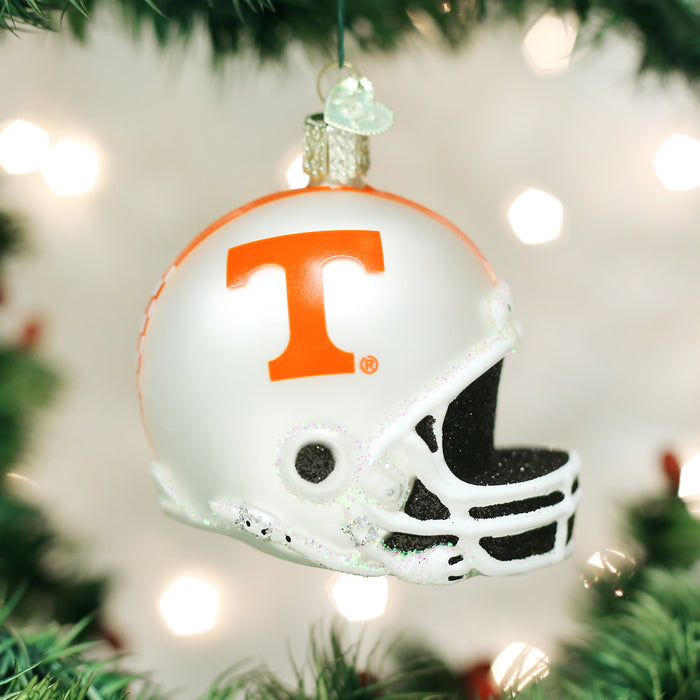 A Christmas tree ornament shaped like a football helmet, decorated with a large orange T on the side, hangs from a green tree branch with white lights in the background.