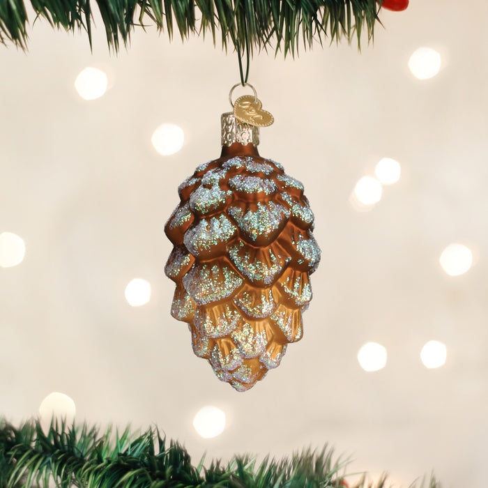 A glittery pine cone-shaped ornament hangs from a green Christmas tree branch with soft, warm lights glowing in the blurred background.